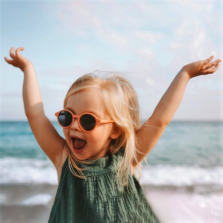 Child girl wearing sunglasses laughing on beach with her arms up